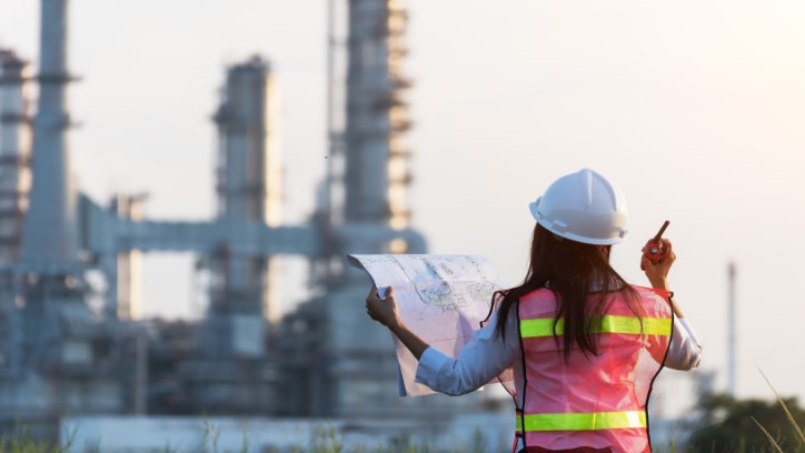 Person wearing a hard hat holding plans and a pencil whilst looking at a large construction in the distance.